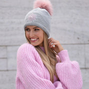 Woman smiling in a pink jumper wearing the SABBOT Jeni bobble hat in grey with pink faux fur pom pom