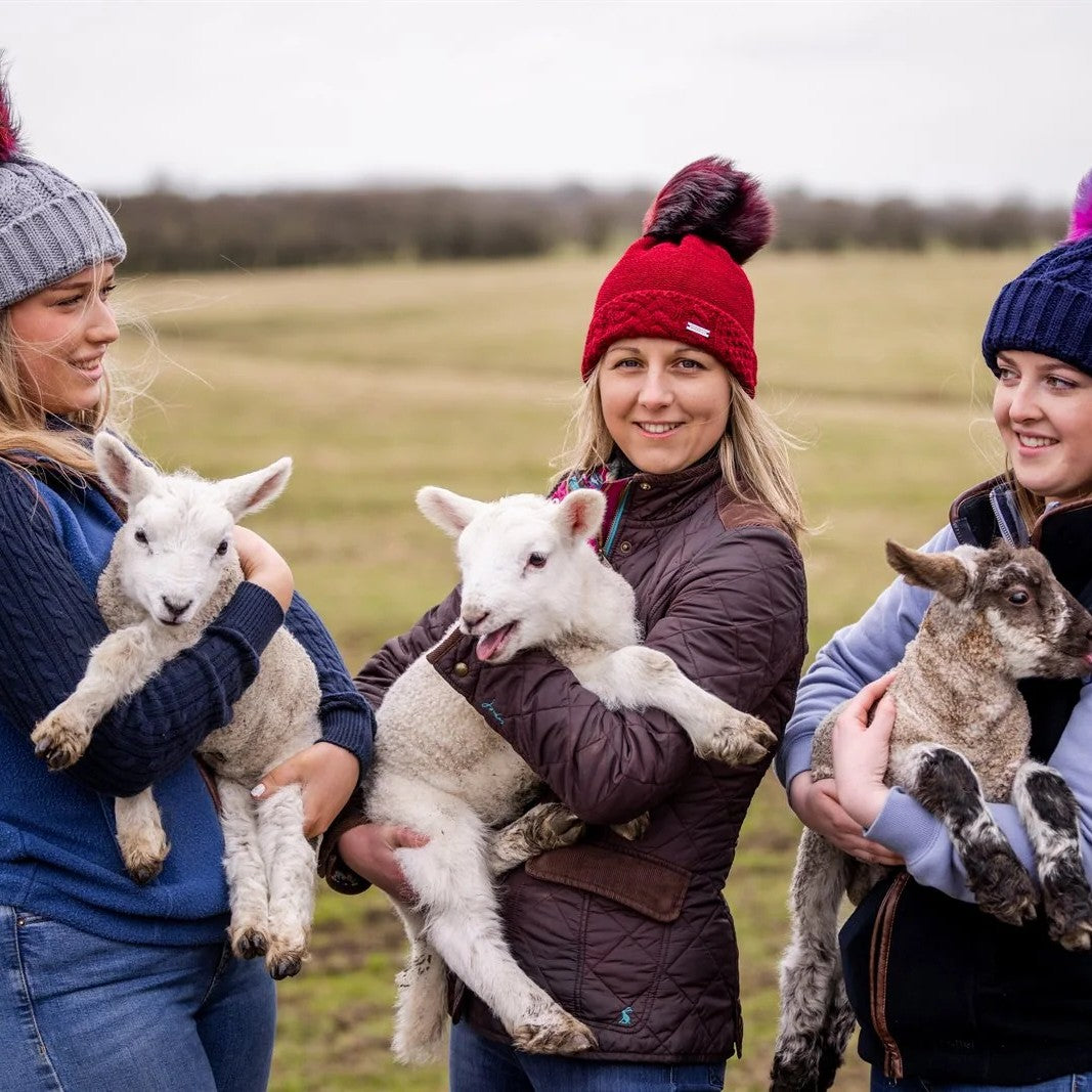 Women cuddling lambs in a field wearing Linda Rainbow and Sigrid bobble hats
