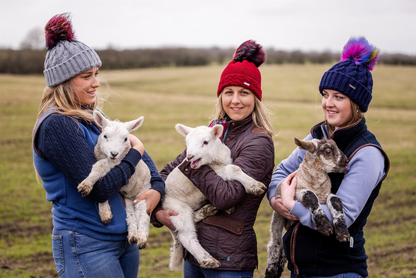 Women cuddling lambs in a field wearing Linda Rainbow and Sigrid bobble hats