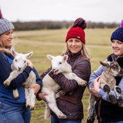 Women cuddling lambs in a field wearing Linda Rainbow and Sigrid bobble hats