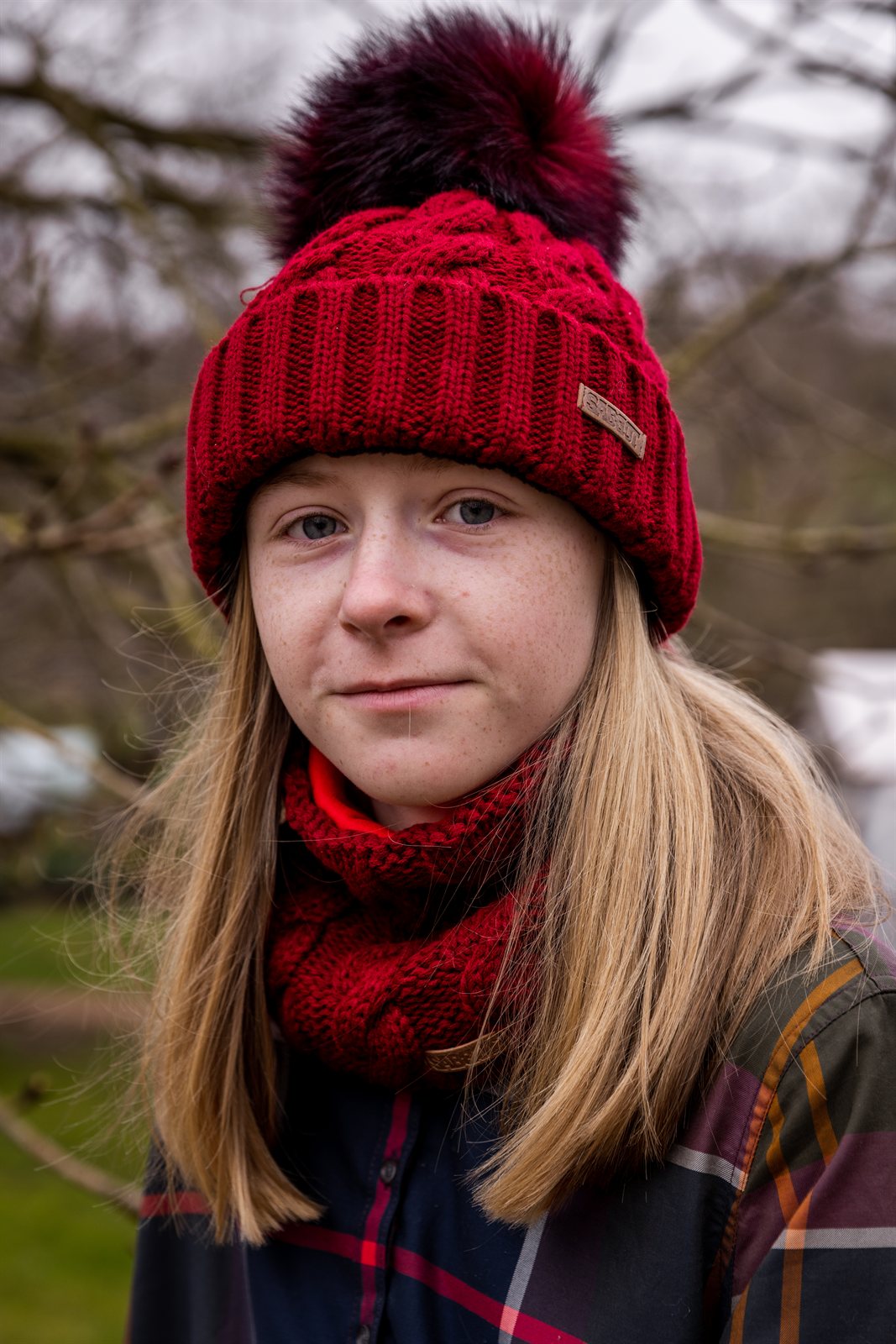 A teenager in tartan in a winter orchard wearing the Linda Bobble Hat and Linda Snood in Burgundy