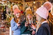 Friends raising a glass at a country pub bar wearing the Linda Bobble Hats in Coral, Mustard and Pink