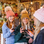 Friends raising a glass at a country pub bar wearing the Linda Bobble Hats in Coral, Mustard and Pink