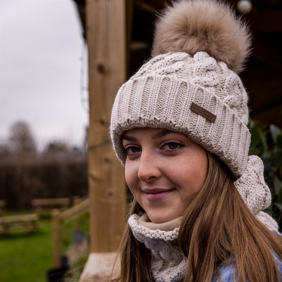 A young woman leaning on a paddock fence wearing the Linda Bobble Hat and Linda Snood in Cream