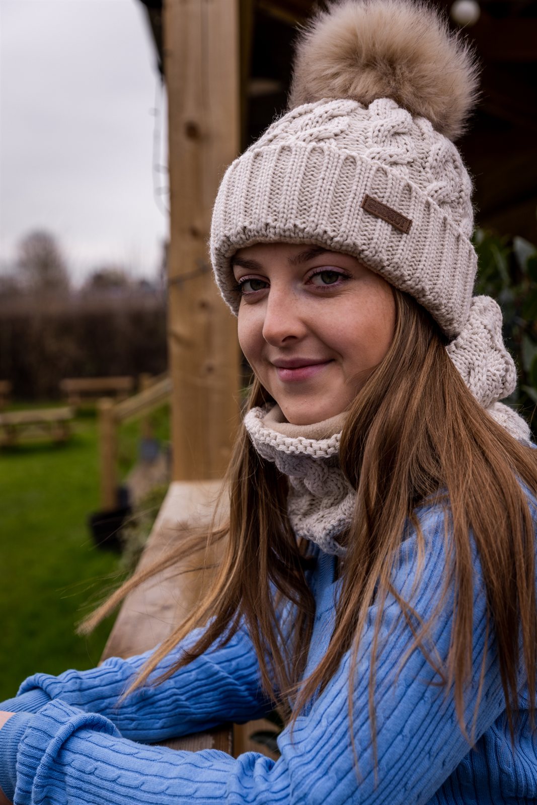 A young woman leaning on a paddock fence wearing the Linda Bobble Hat and Linda Snood in Cream