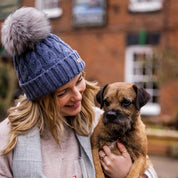 A woman holding a border terrier wearing the Linda Bobble Hat in Denim Blue