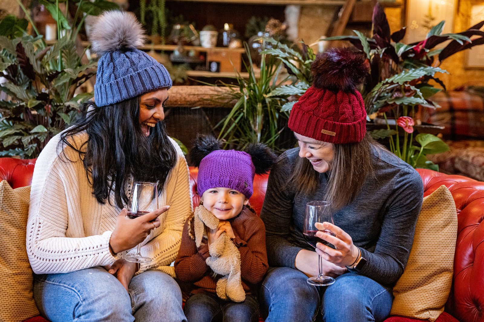 Two women and a toddler on a red leather sofa wearing the Linda Bobble Hats in Denim Blue and Burgundy with a child in a Frankie Childrens Bobble Hat in Purple