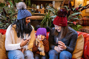 Two women and a toddler on a red leather sofa wearing the Linda Bobble Hats in Denim Blue and Burgundy with a child in a Frankie Childrens Bobble Hat in Purple