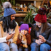 Two women and a toddler on a red leather sofa wearing the Linda Bobble Hats in Denim Blue and Burgundy with a child in a Frankie Childrens Bobble Hat in Purple