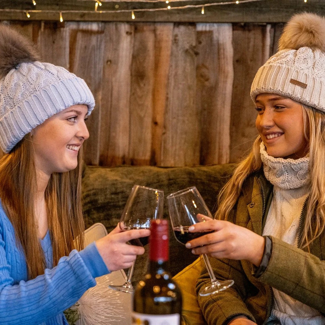 Two friends toasting with red wine on a sofa wearing the Linda Bobble Hats in Light Grey and Cream