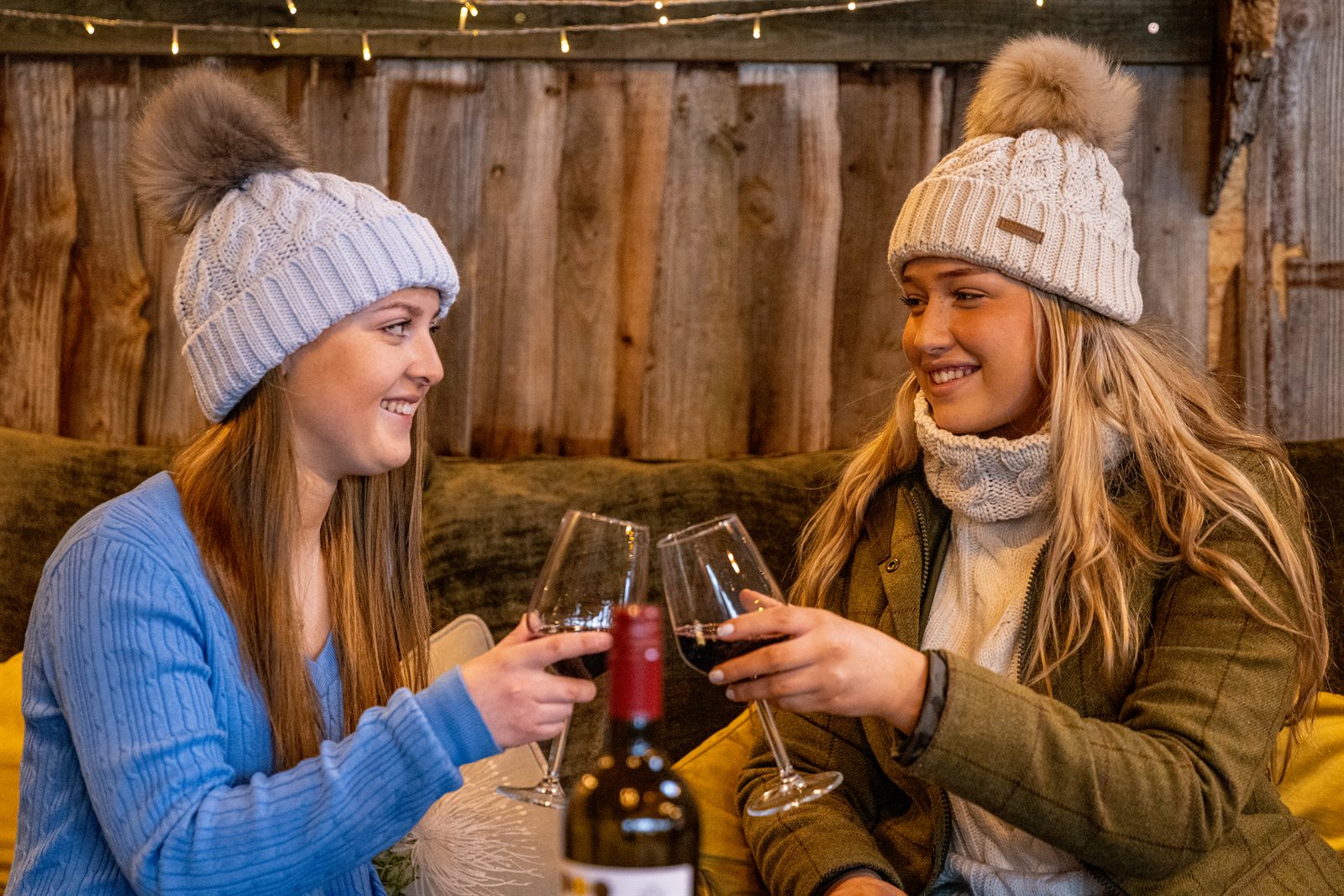 Two friends toasting with red wine on a sofa wearing the Linda Bobble Hats in Light Grey and Cream