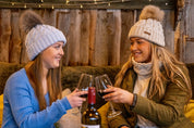Two friends toasting with red wine on a sofa wearing the Linda Bobble Hats in Light Grey and Cream