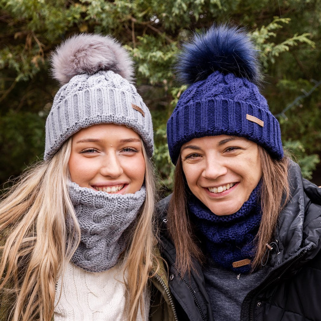 Two friends sharing wine in a winter garden wearing the Linda Bobble Hats and Snoods in Light Grey and Navy Blue