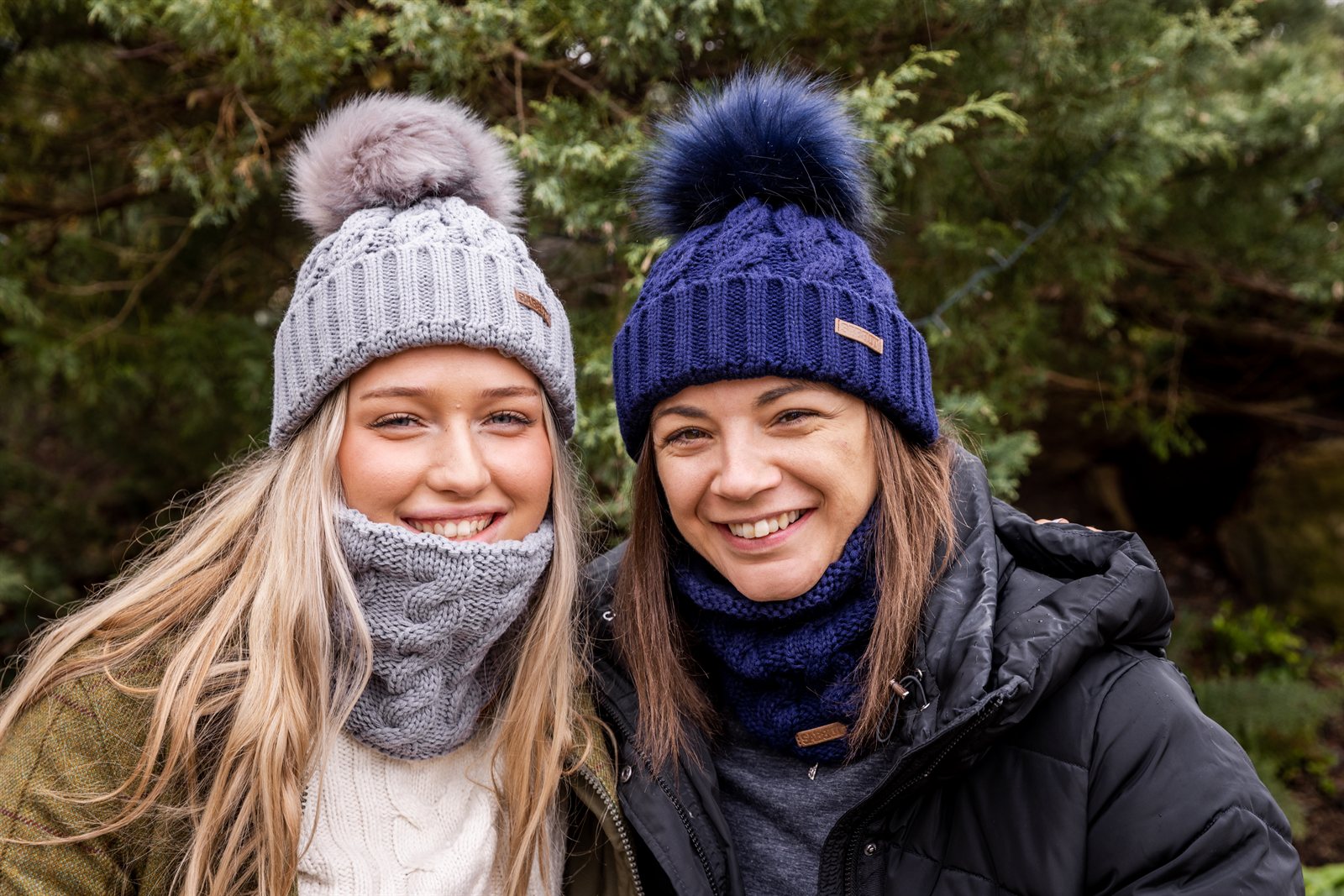 Two friends sharing wine in a winter garden wearing the Linda Bobble Hats and Snoods in Light Grey and Navy Blue