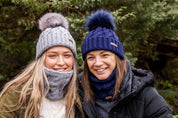 Two friends sharing wine in a winter garden wearing the Linda Bobble Hats and Snoods in Light Grey and Navy Blue
