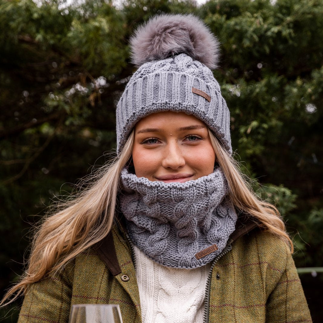 A woman with red wine in a winter garden wearing the Linda Bobble Hat and Linda Snood in Light Grey