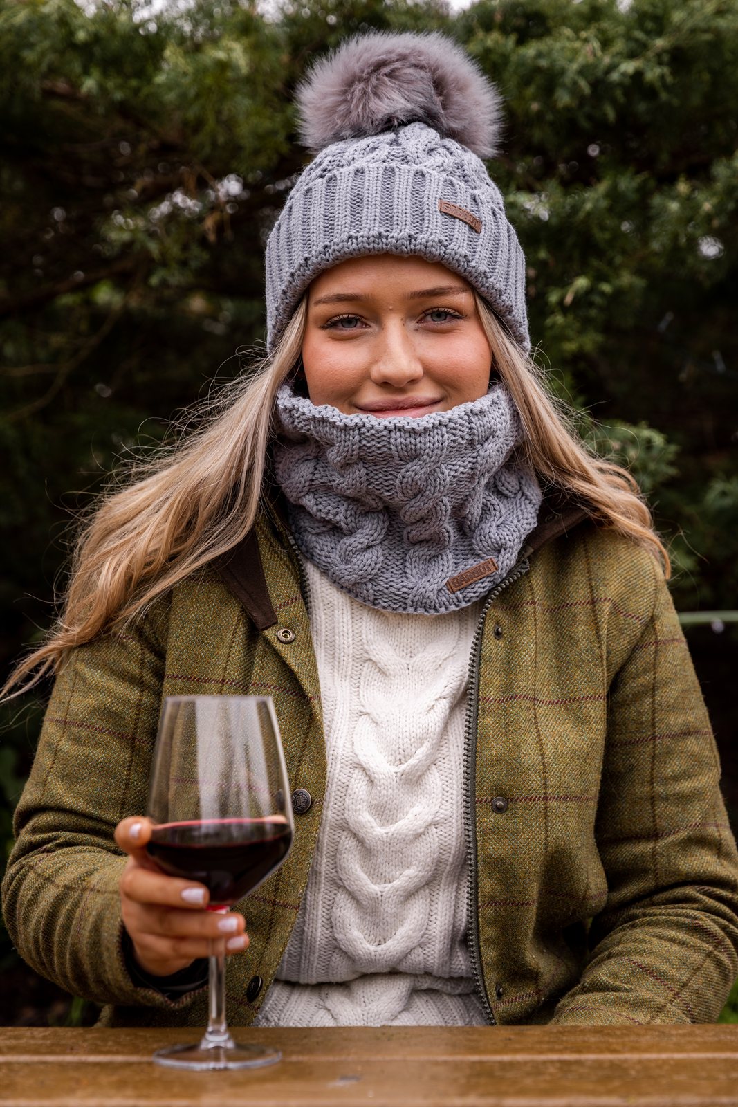 A woman with red wine in a winter garden wearing the Linda Bobble Hat and Linda Snood in Light Grey