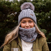 A woman with red wine in a winter garden wearing the Linda Bobble Hat and Linda Snood in Light Grey