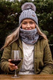 A woman with red wine in a winter garden wearing the Linda Bobble Hat and Linda Snood in Light Grey