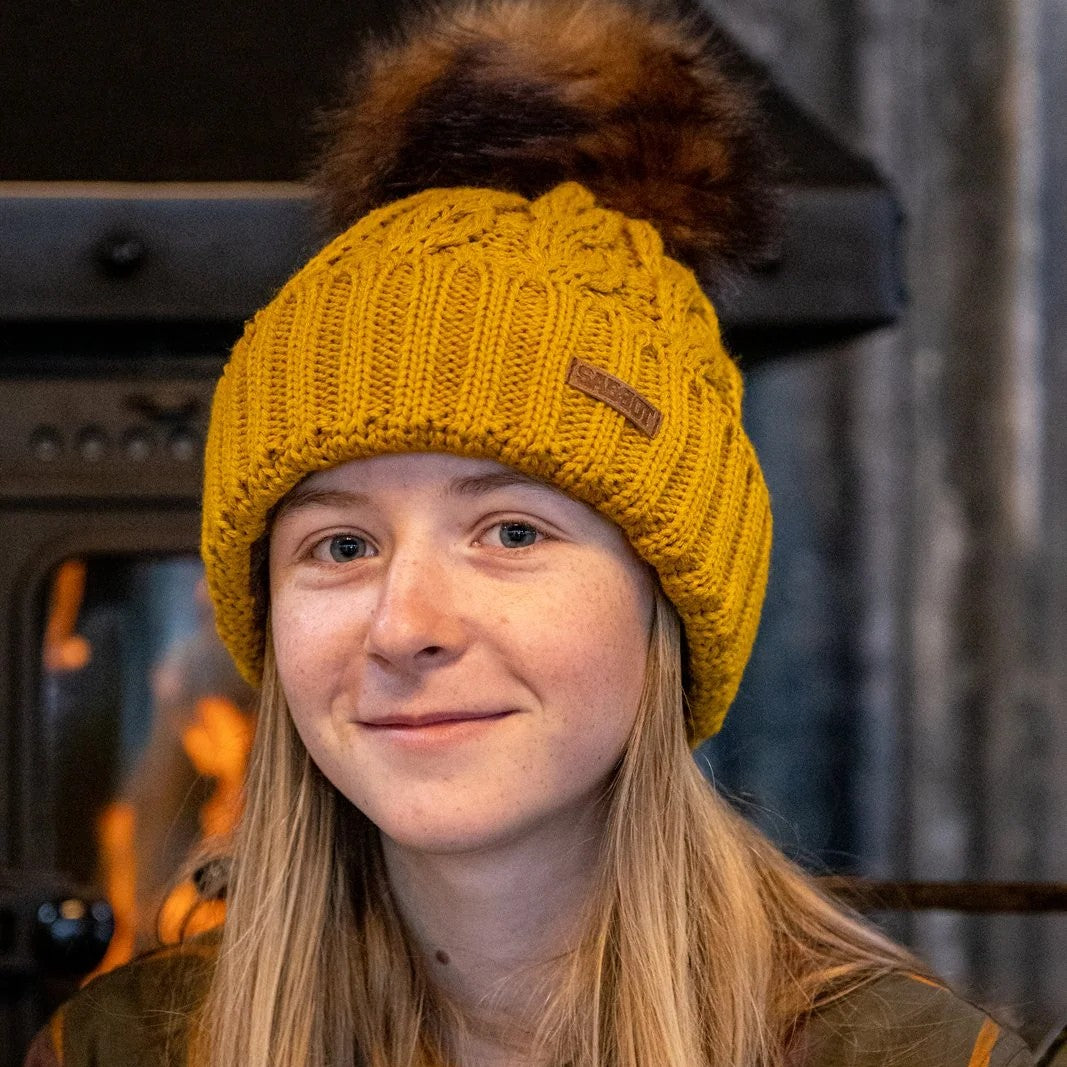 Two women by a log burner wearing the Linda Bobble Hat in Mustard