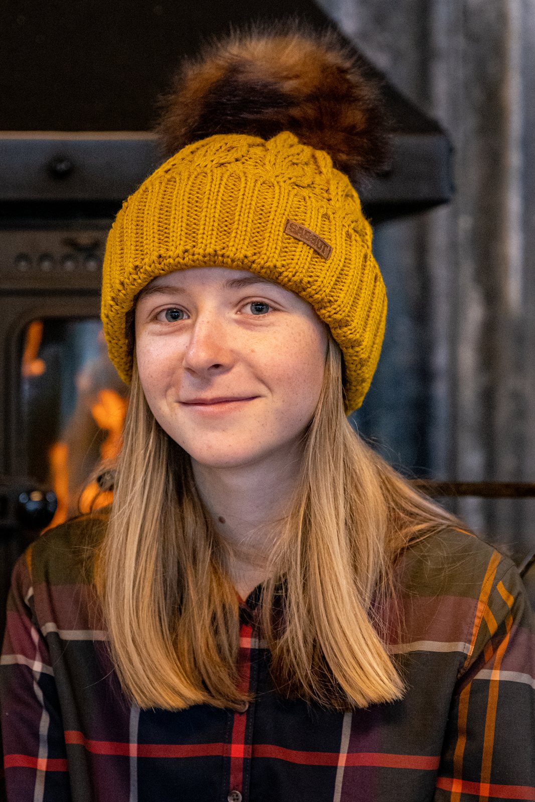 Two women by a log burner wearing the Linda Bobble Hat in Mustard