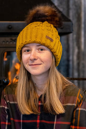 Two women by a log burner wearing the Linda Bobble Hat in Mustard