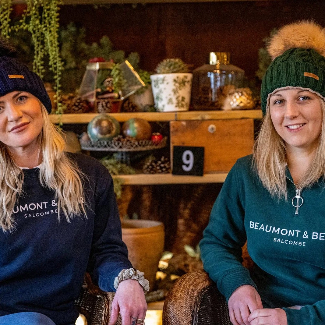 Two friends chatting in a cosy plant-filled bar wearing the Linda Bobble Hat in Navy Blue and Forest