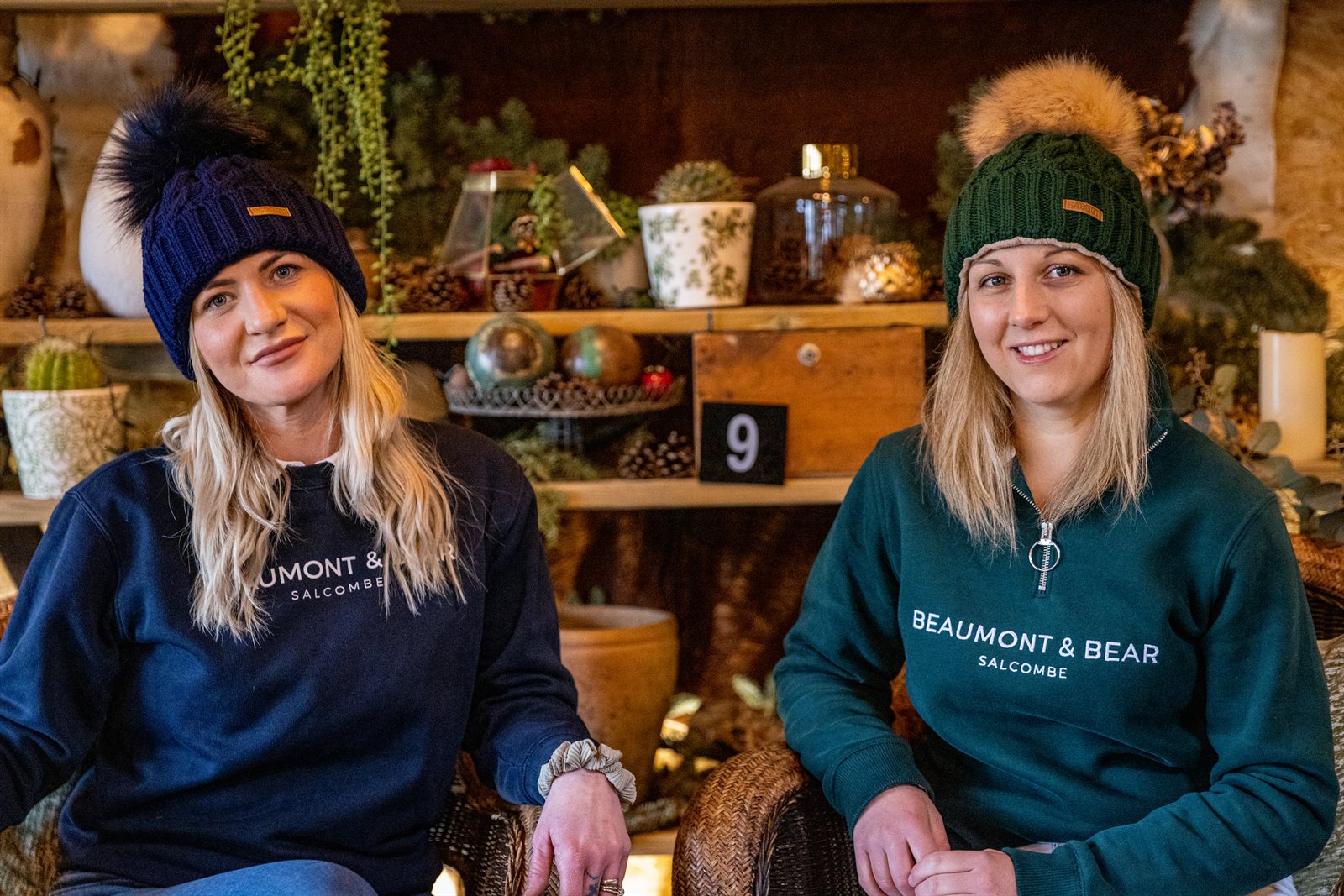 Two friends chatting in a cosy plant-filled bar wearing the Linda Bobble Hat in Navy Blue and Forest