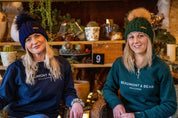 Two friends chatting in a cosy plant-filled bar wearing the Linda Bobble Hat in Navy Blue and Forest