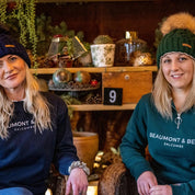 Two friends chatting in a cosy plant-filled bar wearing the Linda Bobble Hat in Navy Blue and Forest