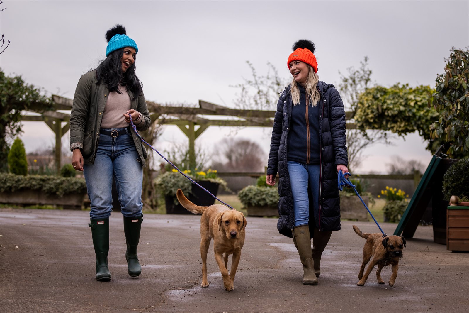 Three friends out walking their dogs wearing the Linda Neon Bobble Hats in Neon Blue and Neon Orange