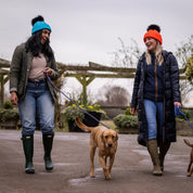 Three friends out walking their dogs wearing the Linda Neon Bobble Hats in Neon Blue and Neon Orange