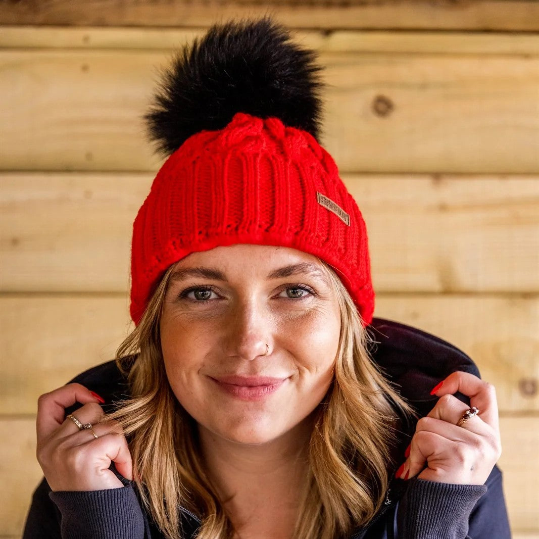 A woman pulling up her collar against a timber wall wearing the Linda Neon Bobble Hat in Neon Red
