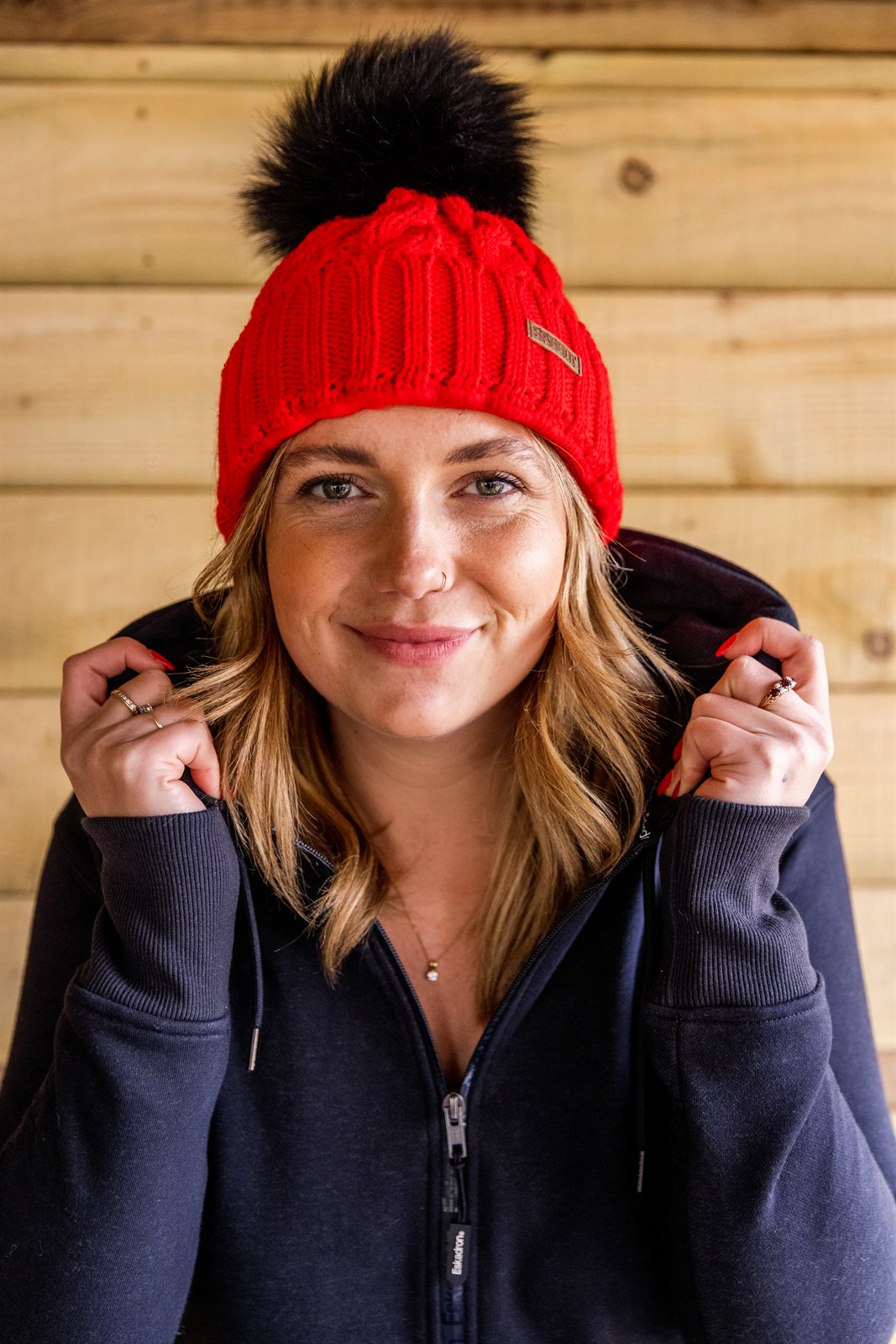 A woman pulling up her collar against a timber wall wearing the Linda Neon Bobble Hat in Neon Red