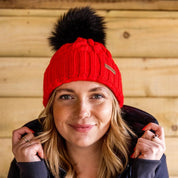 A woman pulling up her collar against a timber wall wearing the Linda Neon Bobble Hat in Neon Red