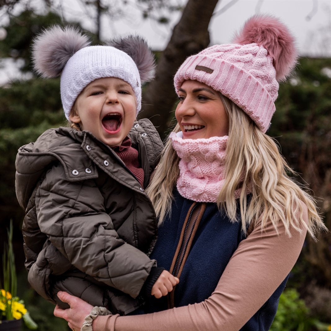 A mum laughing with her toddler outdoors wearing the Linda Bobble Hat and Snood in Pink with a Frankie Childrens Bobble Hat in White