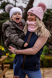 A mum laughing with her toddler outdoors wearing the Linda Bobble Hat and Snood in Pink with a Frankie Childrens Bobble Hat in White