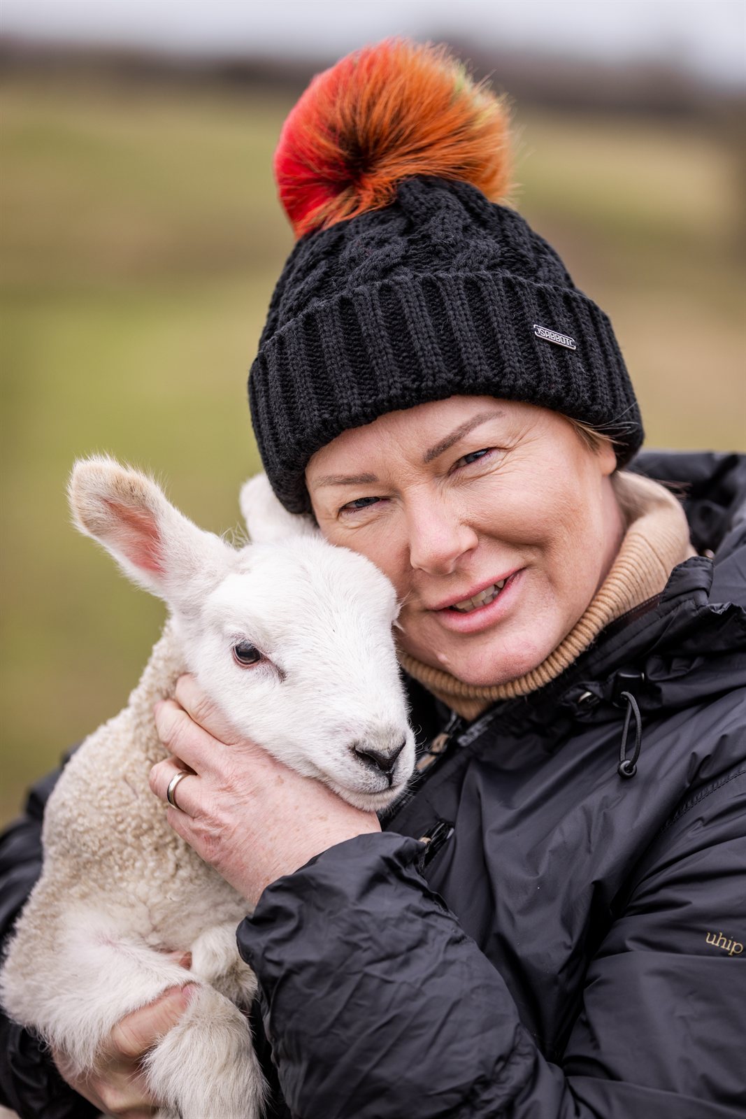 Women cuddling lambs in a field wearing Linda Rainbow Bobble Hat in Black