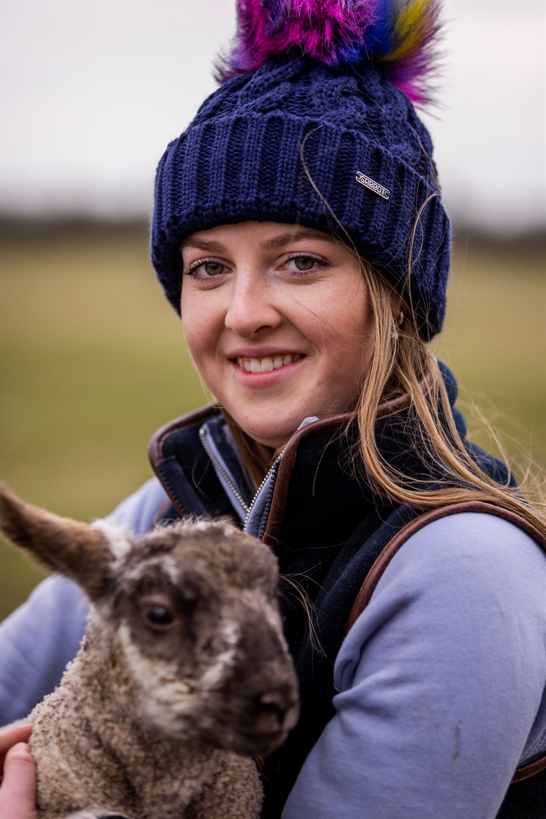 Women cuddling lambs in a field wearing Linda Rainbow Bobble Hat in Navy Blue