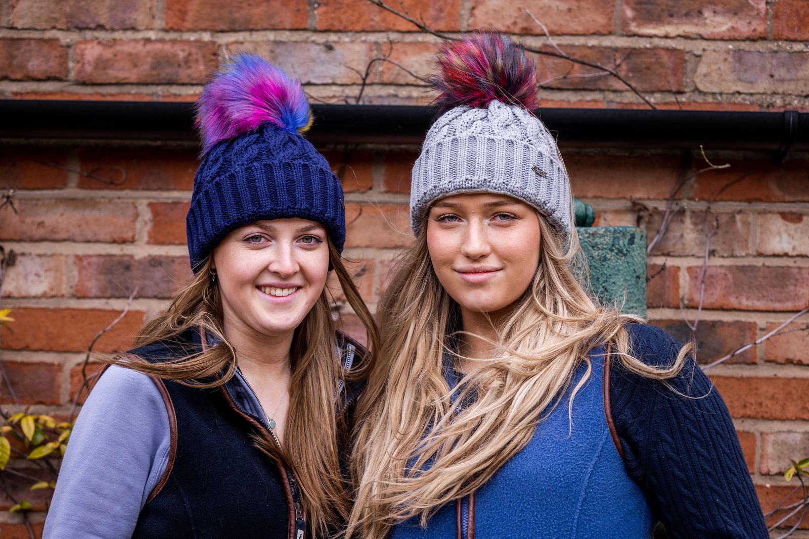 Friends in rainbow pom-pom hats by an old brick wall wearing the Linda Rainbow Bobble Hats in Navy Blue and Light Grey
