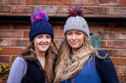 Friends in rainbow pom-pom hats by an old brick wall wearing the Linda Rainbow Bobble Hats in Navy Blue and Light Grey