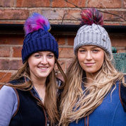 Friends in rainbow pom-pom hats by an old brick wall wearing the Linda Rainbow Bobble Hats in Navy Blue and Light Grey