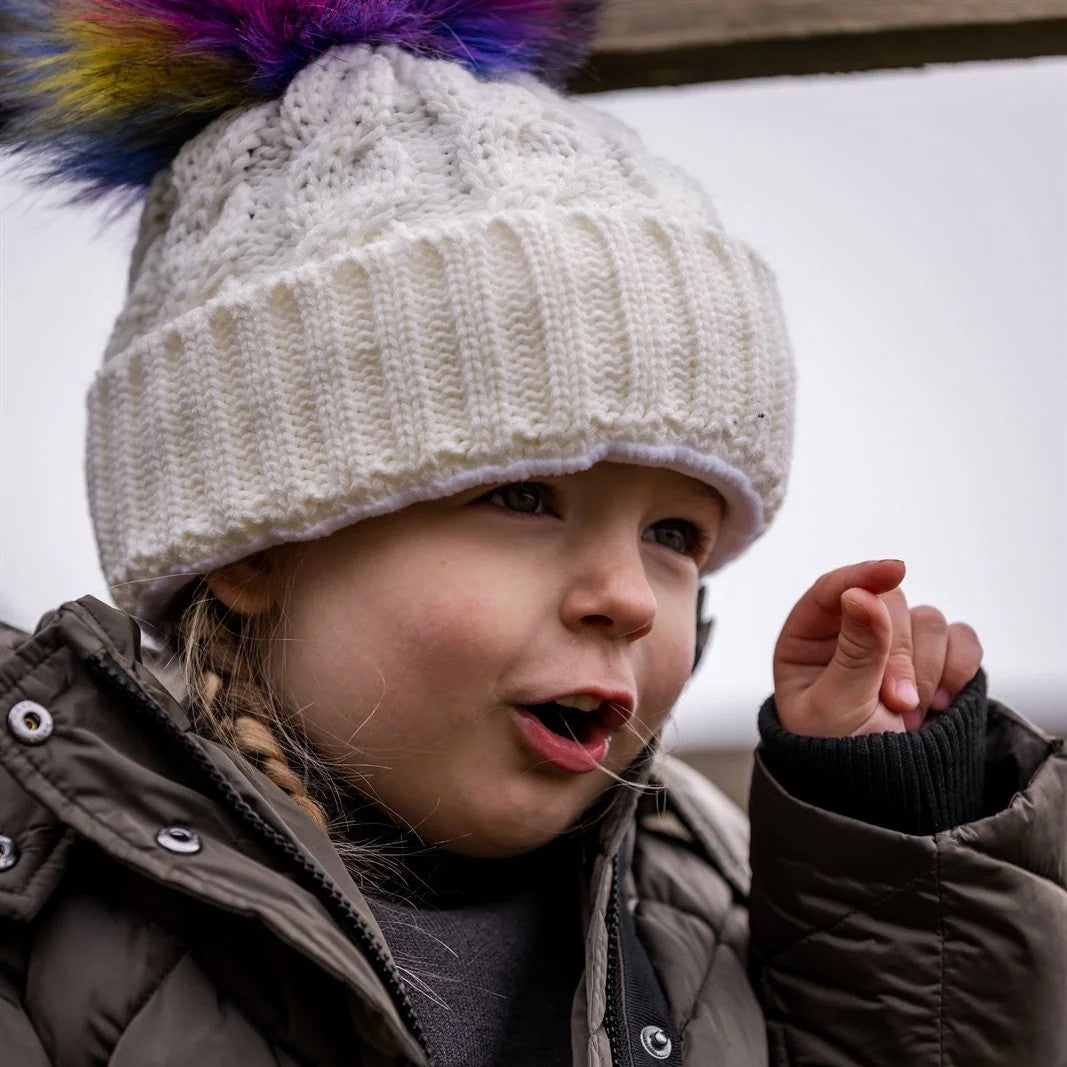 A young child in a white rainbow pom-pom hat wearing the Linda Rainbow Bobble Hat in White