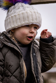A young child in a white rainbow pom-pom hat wearing the Linda Rainbow Bobble Hat in White