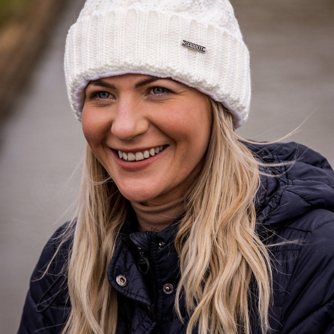 Friends in rainbow pom-pom hats by an old brick wall wearing the Linda Rainbow Bobble Hat in White