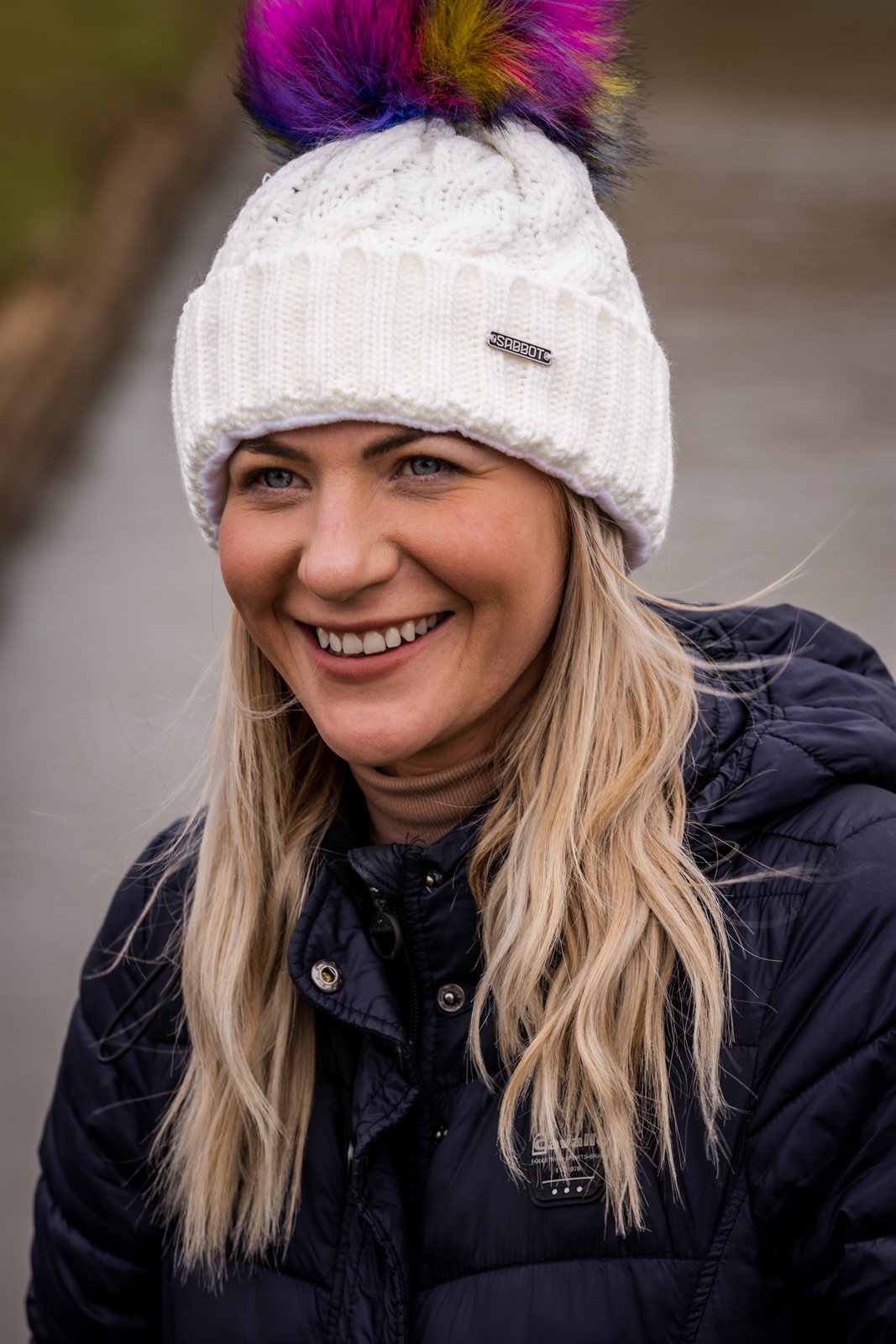 Friends in rainbow pom-pom hats by an old brick wall wearing the Linda Rainbow Bobble Hat in White
