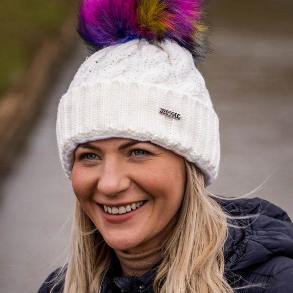 Friends in rainbow pom-pom hats by an old brick wall wearing the Linda Rainbow Bobble Hat in White