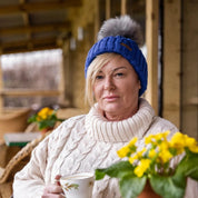 A woman drinking tea on a cabin porch wearing the Linda Bobble Hat in Royal