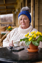 A woman drinking tea on a cabin porch wearing the Linda Bobble Hat in Royal