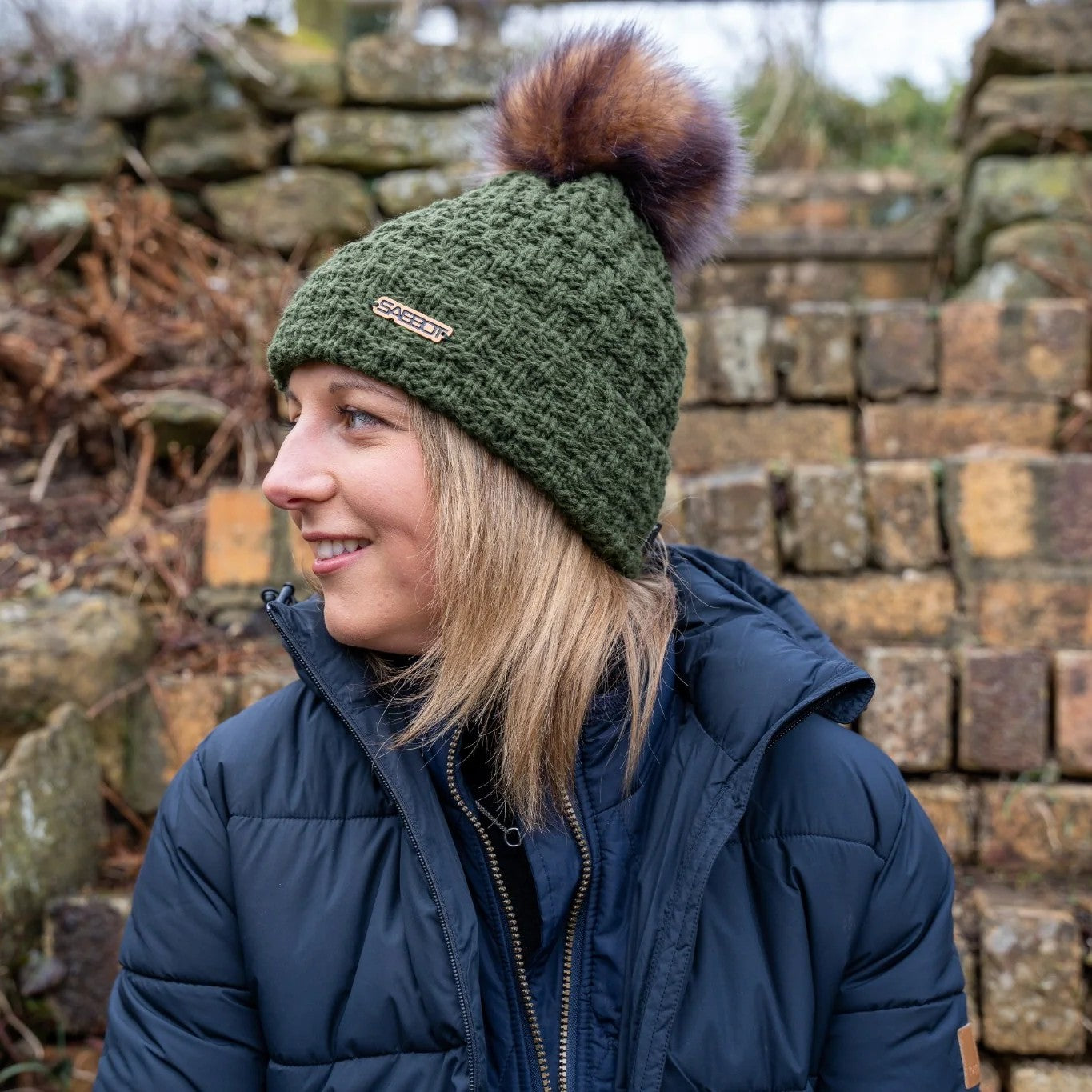 A woman by an old stone wall wearing the Petra Fold Bobble Hat in Olive with faux fur pom pom